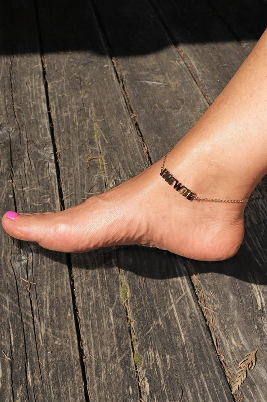 Ankle bracelet on a beautiful females foot with pink nail polish on a wooden surface. The anklet has a "Hotwife" logo / charm on it.