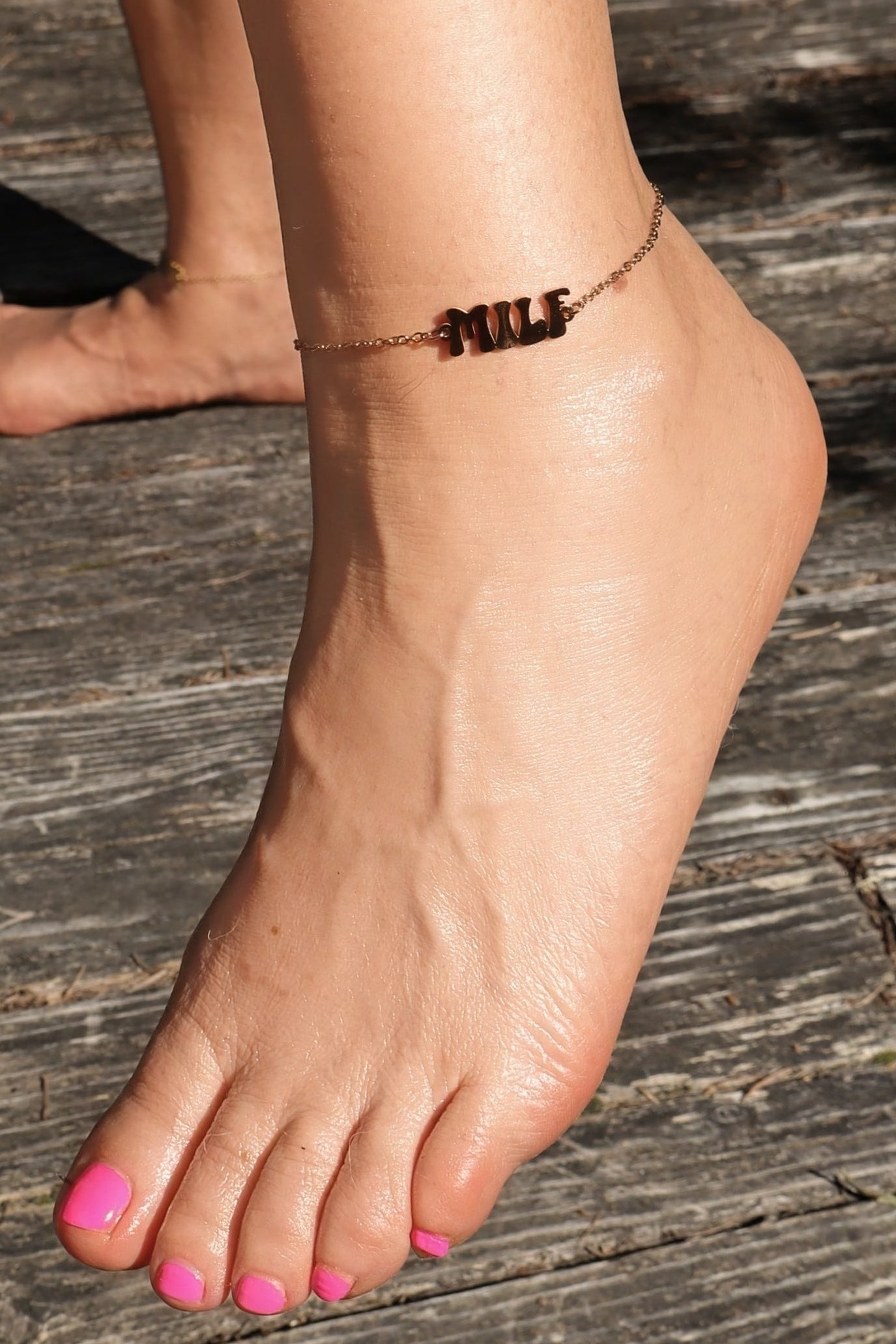 Close-up of a beautiful female's foot wearing a gold anklet with "MILF" logo on a wooden surface with bright pink pedicure.