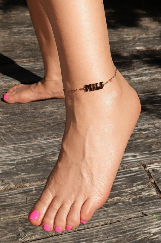 Close-up of a beautiful female's foot wearing a gold anklet with "MILF" logo on a wooden surface with bright pink pedicure.