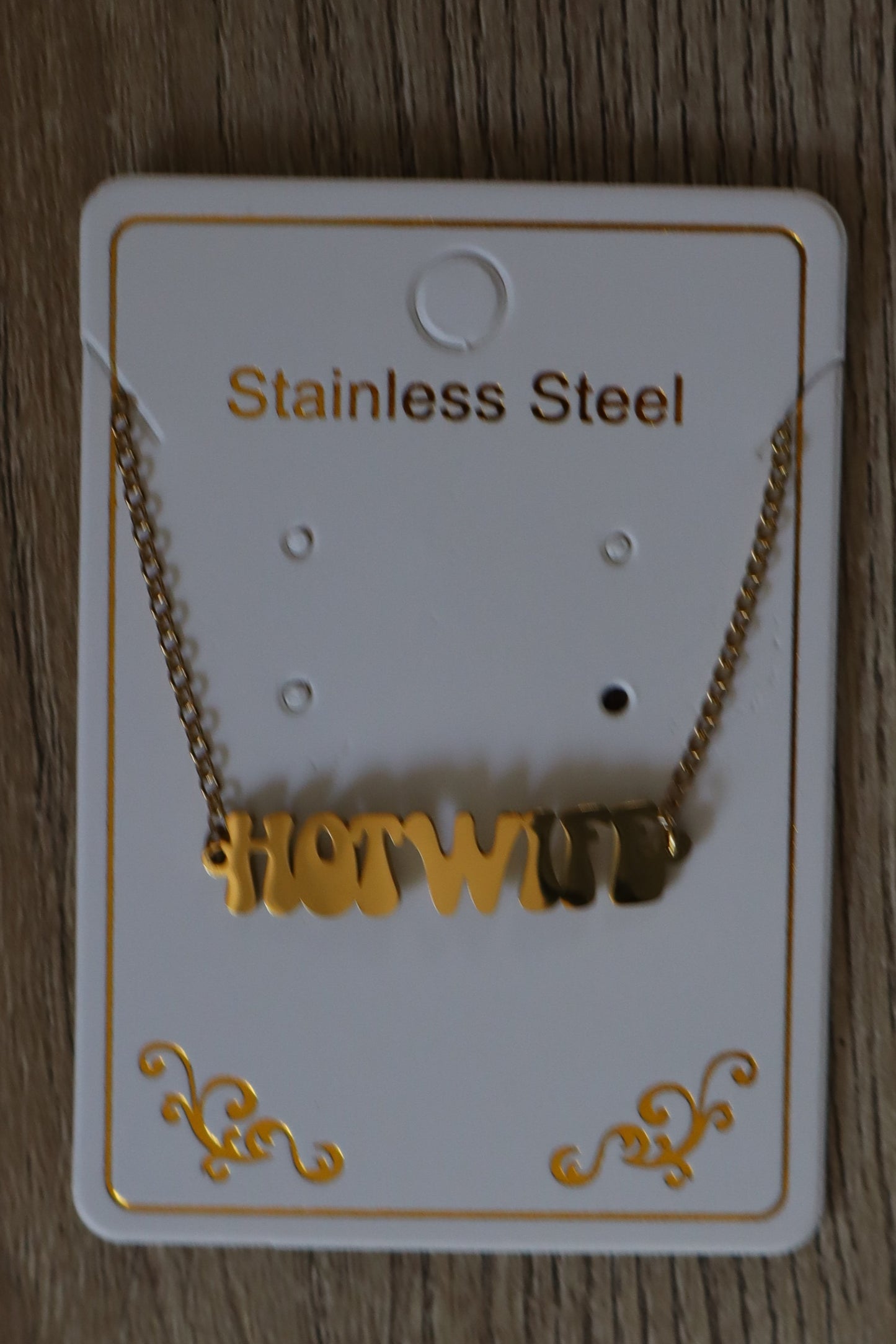 Gold anklet / bracelet with a "HOTWIFE" logo / charm on a white card on a wooden surface.